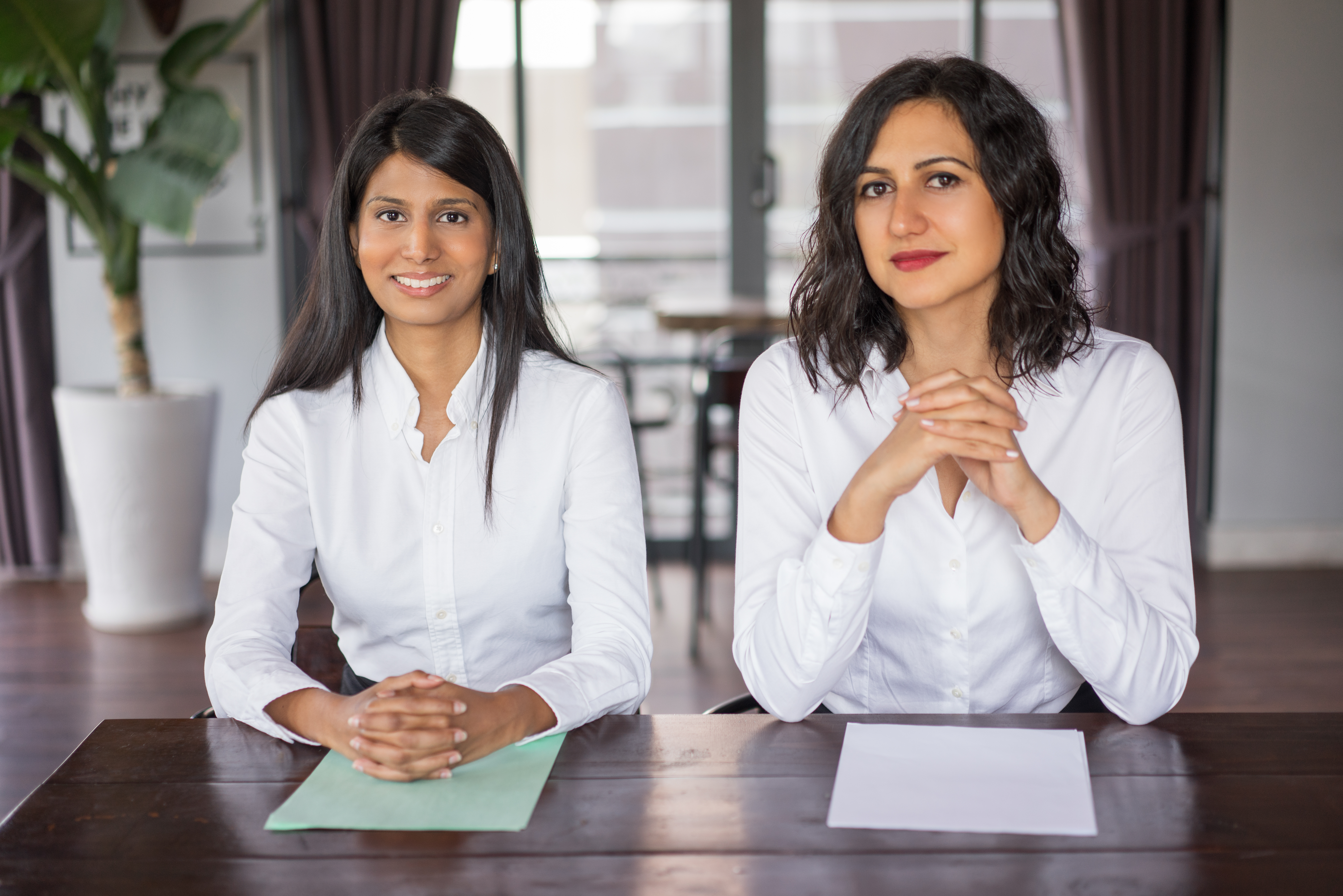 Two positive female workers sitting at desk in modern office. They are looking at camera. Female employees concept. Front view.